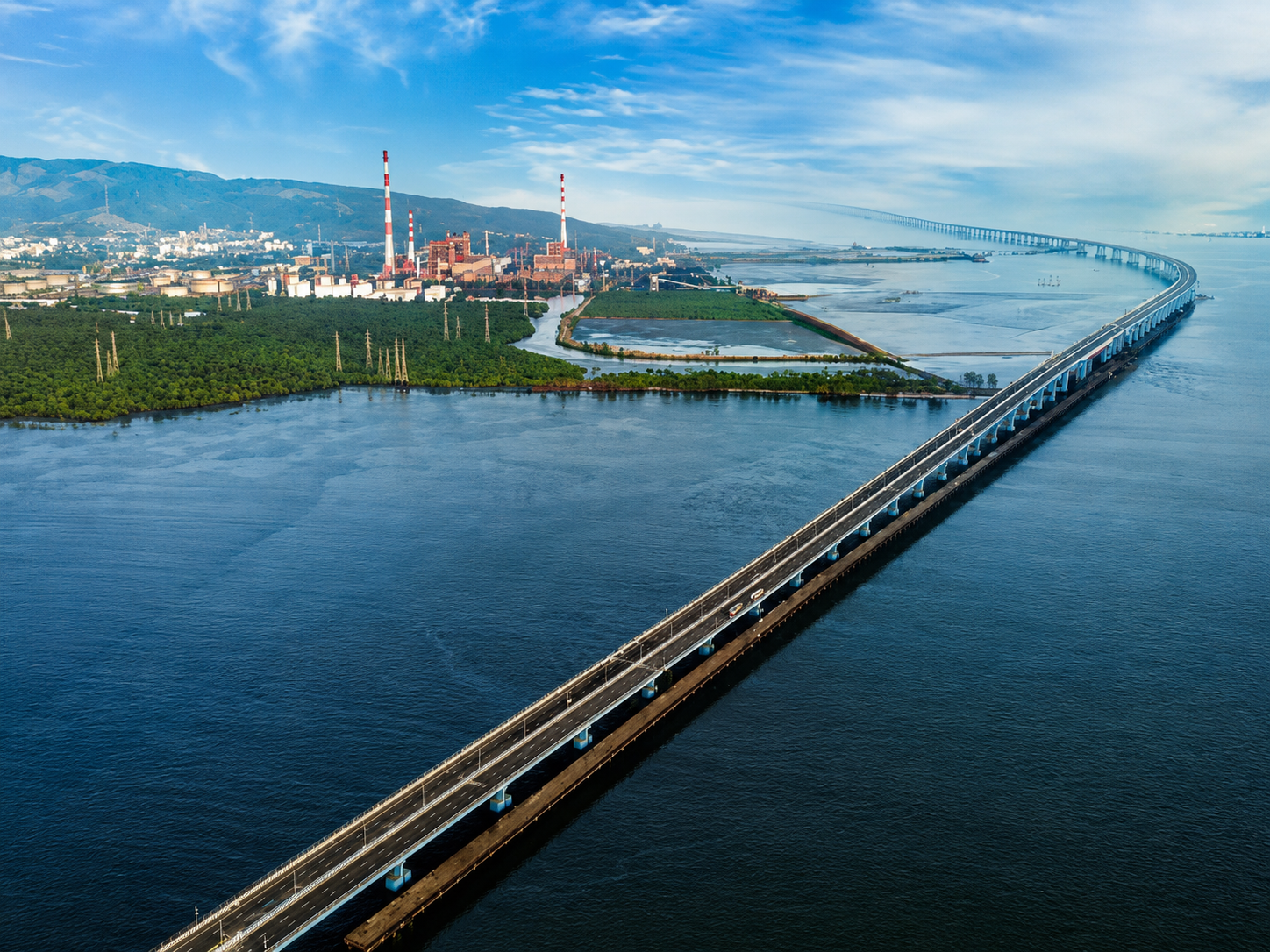 Aerial view of the Mumbai–Alibaug coastal corridor, featuring the Mumbai Trans Harbour Link (Atal Setu) stretching across the Arabian Sea, with mangroves, coastal infrastructure, industrial facilities, and distant hills visible in the background.